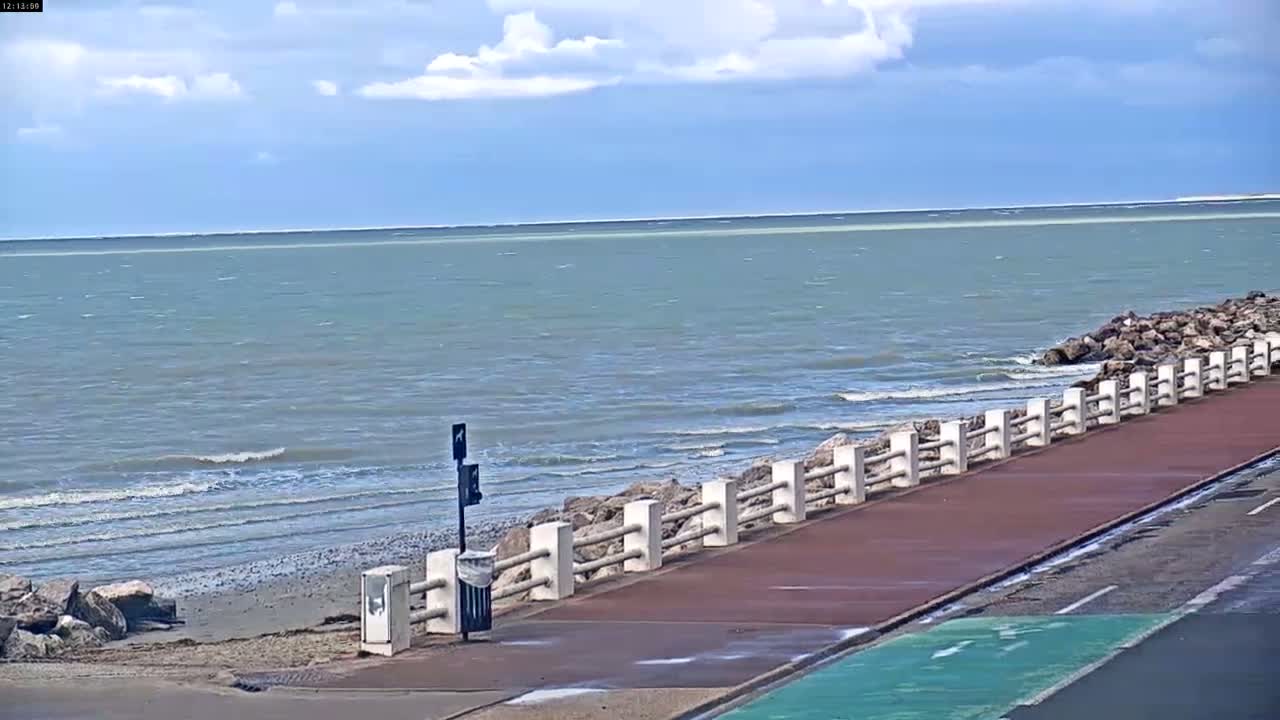A paved walkway with a low stone fence separates a calm, grey-green sea with small waves from a shoreline of rocks and sand under a partly cloudy sky.