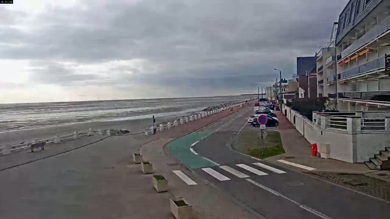 A seaside road with parked cars and apartment buildings curves alongside a beach under a cloudy sky.