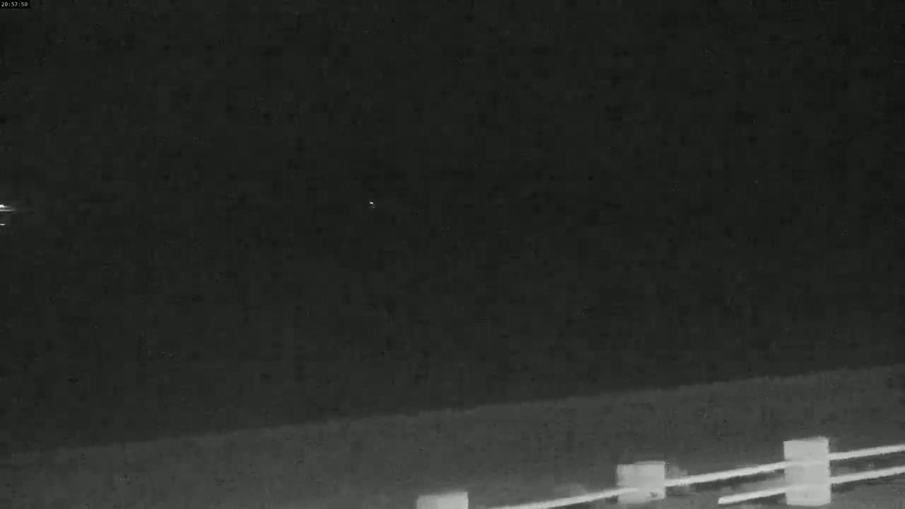 A nighttime, low-light view of a dark, flat landscape with a low fence in the bottom right corner.