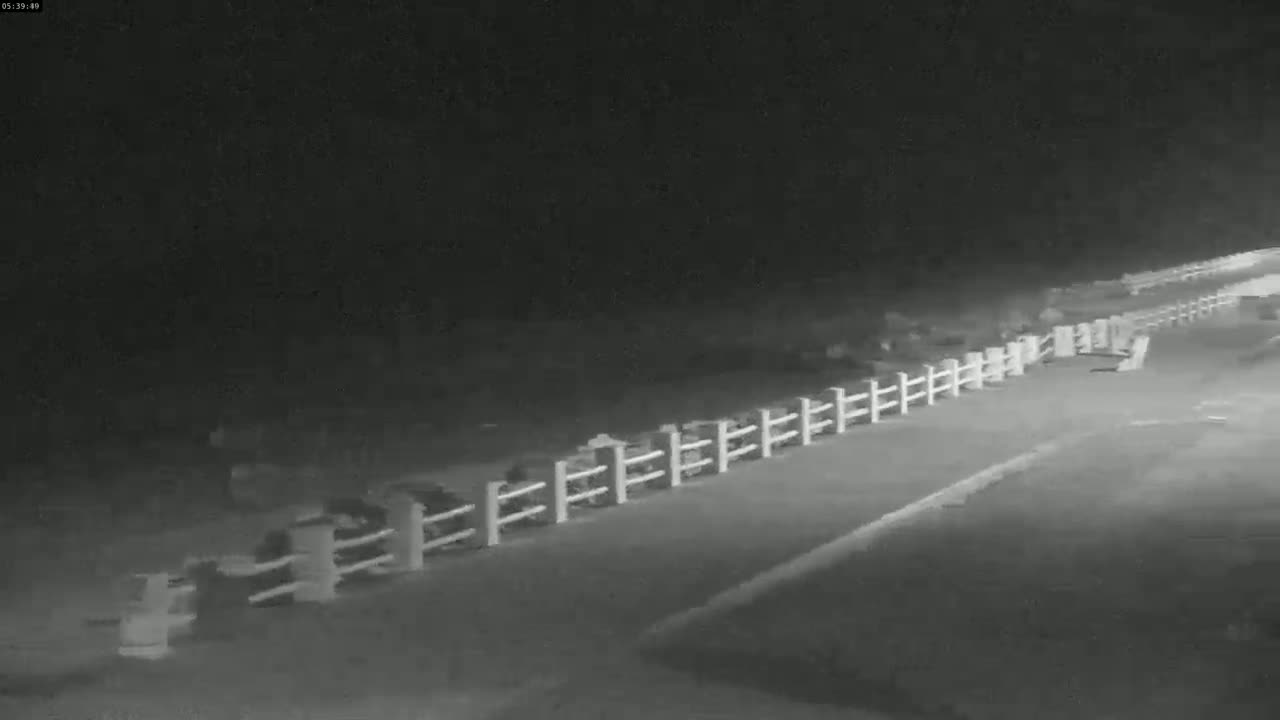 A nighttime, low-light image shows a long white fence running alongside a paved road, with indistinct objects visible behind the fence.