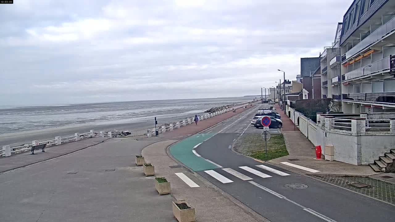 A paved walkway and road beside a beach with low tide under a cloudy sky, next to a row of buildings.