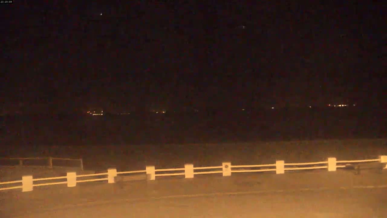 A nighttime view shows a white railing in the foreground, a dark expanse of land in the middle ground, and distant lights on the horizon under a clear, dark sky.