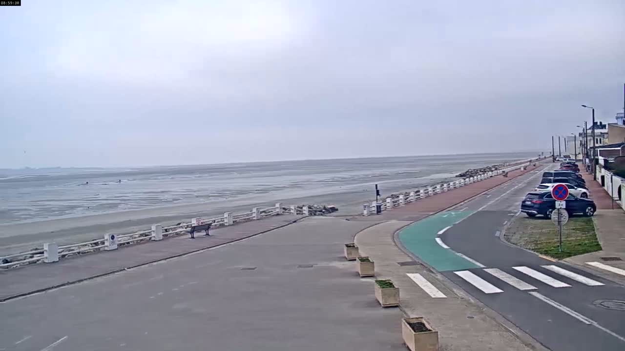 A mostly empty seaside promenade with a few parked cars and a white fence separating it from a wide expanse of mudflats under a cloudy sky.