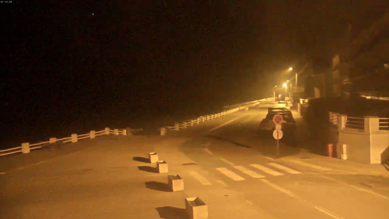 A nighttime coastal road scene is illuminated by streetlights under hazy or foggy conditions, showing a parked car and a low white fence bordering the ocean.