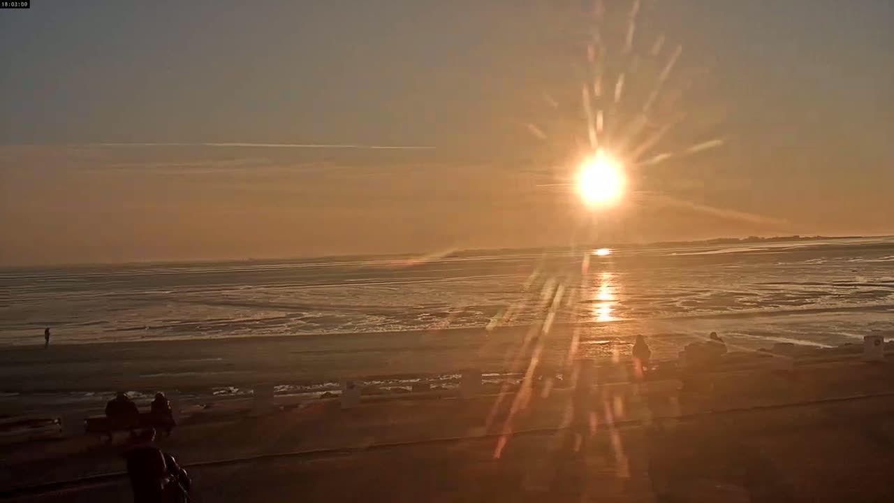 A low-lying sun sets over a wide expanse of calm, sandy beach and ocean under a clear, orange-hued sky.
