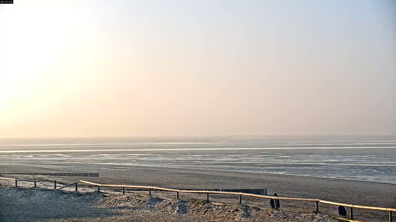 A hazy sunrise illuminates a wide, flat beach with a low wooden fence in the foreground, overlooking a tidal flat.
