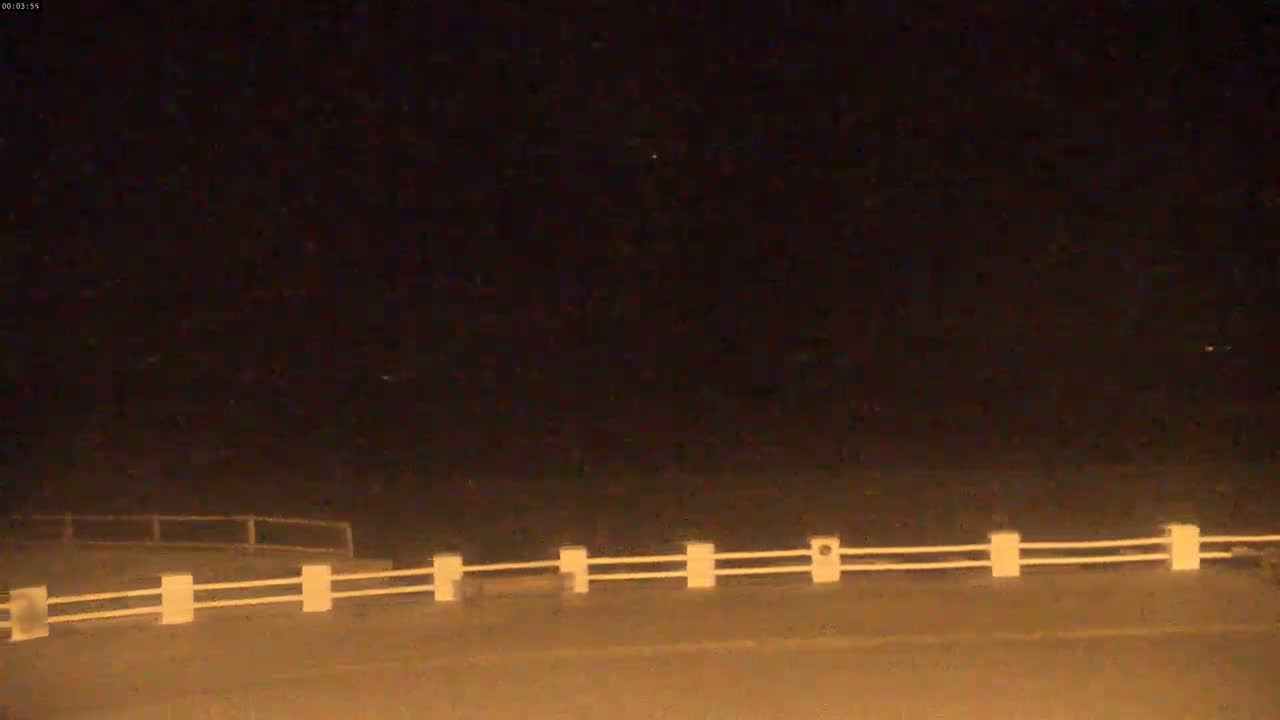 A low-light nighttime image shows a white railing along a dark roadway, under a dark sky.