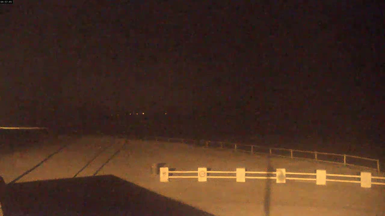 A dark, nighttime scene shows a paved area with a low white railing, dimly lit by a few distant lights.
