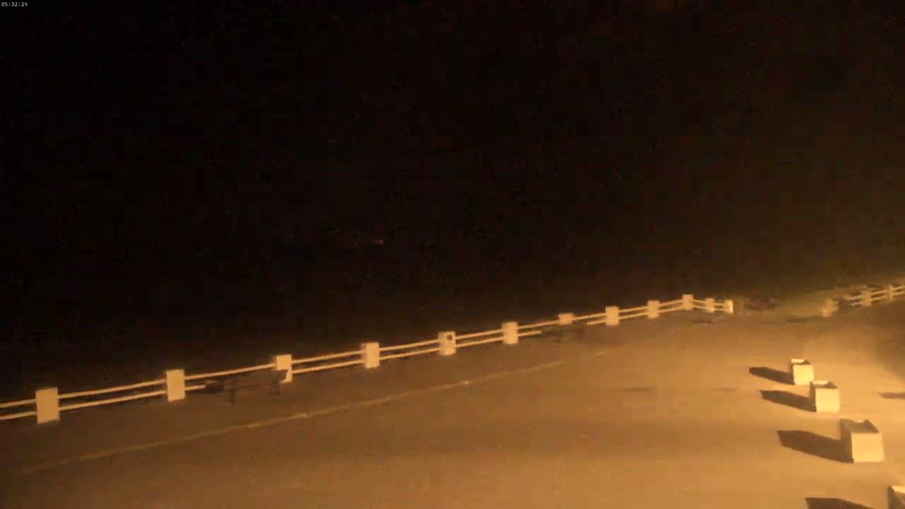 A nighttime, dimly lit outdoor scene shows a low white fence running along the edge of a paved area, with several dark objects near the fence and a few light-colored square objects in the lower right.