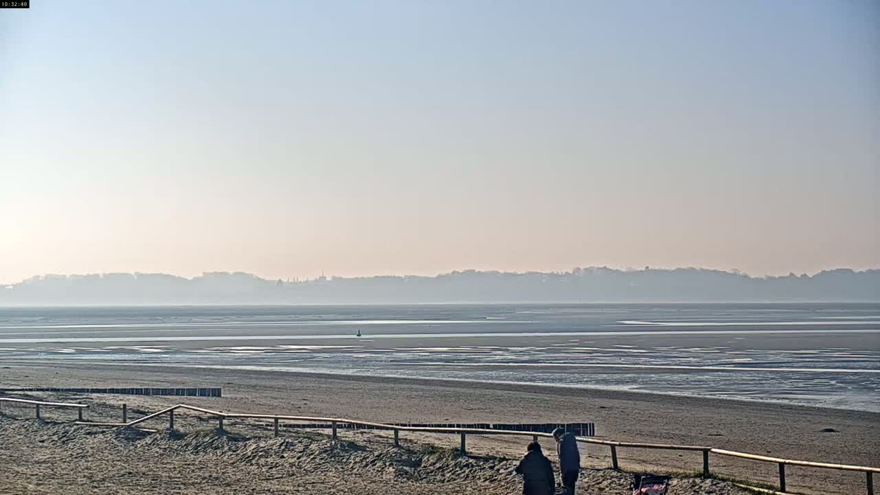 Two people stand near a wooden fence on a sandy beach overlooking a hazy expanse of tidal flats and a distant shoreline under a clear, pale sky.