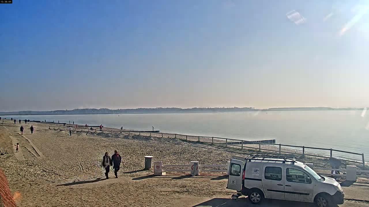 A sandy beach with a calm body of water, a few people walking, and a white van parked nearby under a clear, sunny sky.