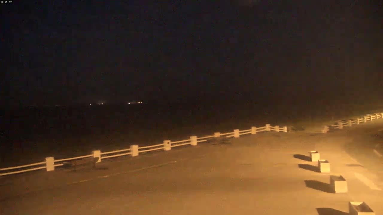 A nighttime view of a paved area with a low white fence separating it from a dark body of water, under a clear, dark sky.