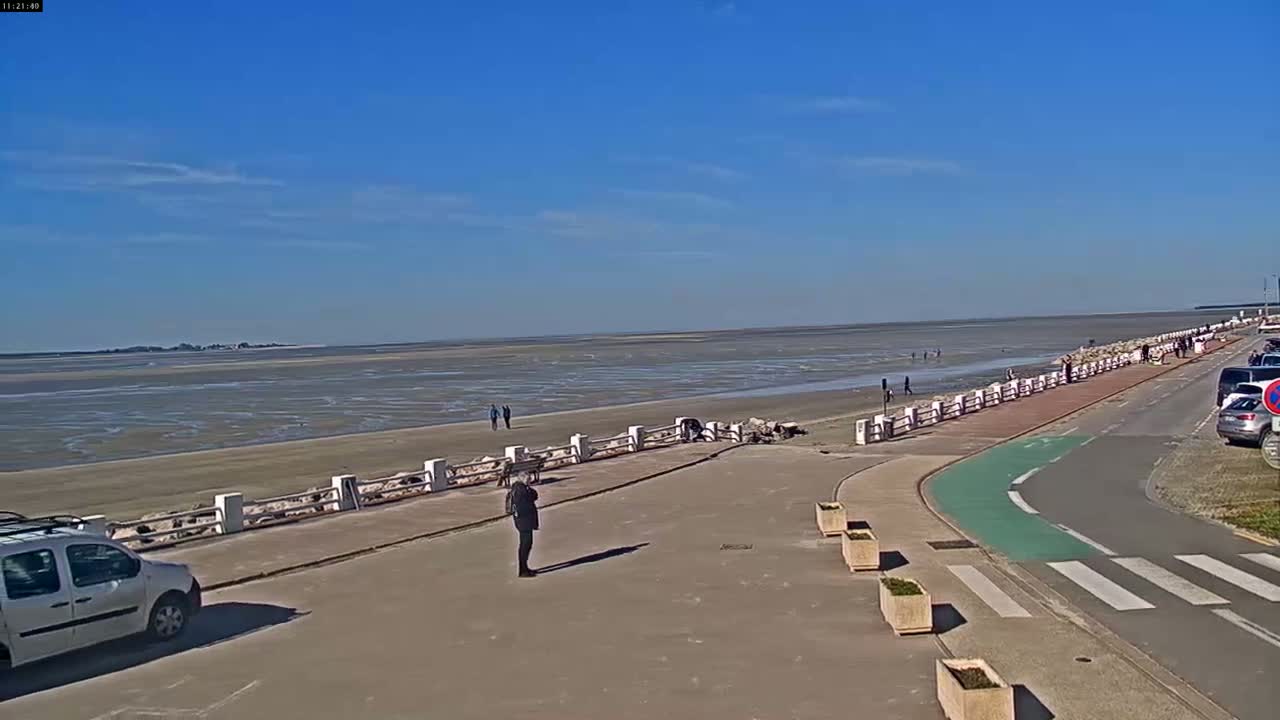 A wide, paved waterfront path with a few people walking along it overlooks a mostly exposed, flat sandy beach and calm, shallow water under a clear, sunny sky.