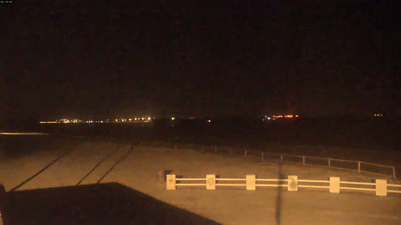 A nighttime view of a flat, dark landscape with a distant line of lights and a short, low white fence in the foreground.