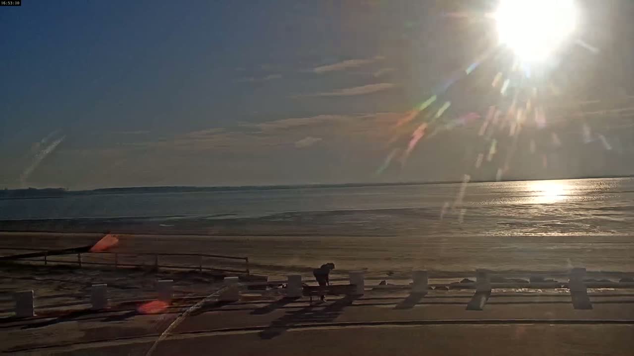 A person stands near a low wall overlooking a calm, sunlit body of water on a mostly clear day.