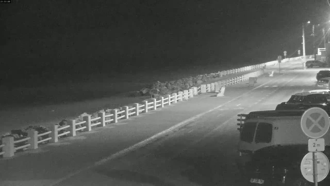 A nighttime view of a road next to a rocky shoreline, separated by a short white fence, with several cars parked along the roadside.