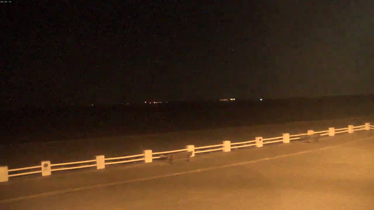 A dark, nighttime scene shows a low white fence running parallel to a road next to a body of water with distant lights visible on the horizon.