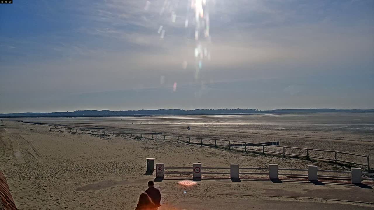 A sandy beach stretches to a distant shoreline under a bright, sunny sky with a few clouds.