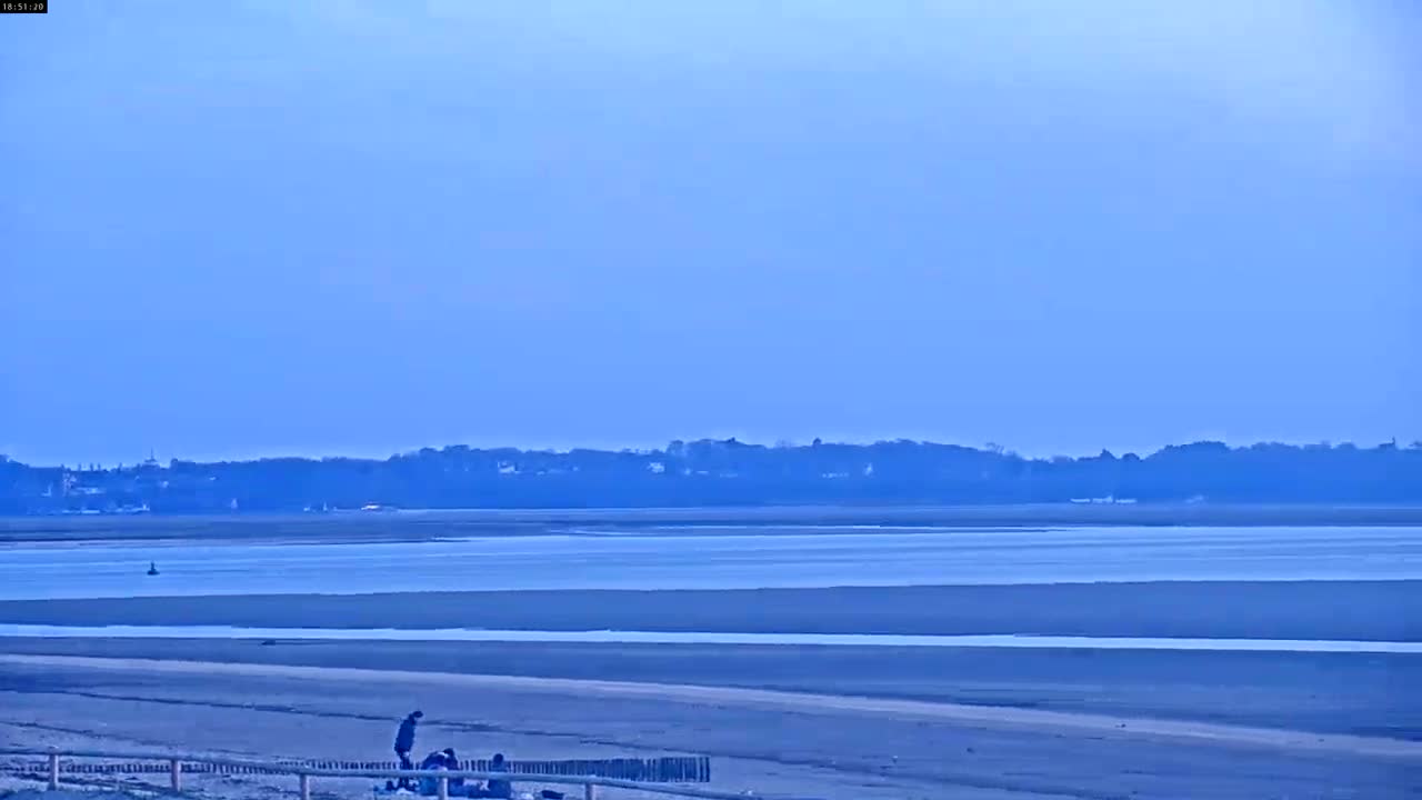 A wide, flat expanse of sandy beach stretches to a calm, light-blue body of water under a clear, pale-blue sky, with a few people visible near the shoreline and a distant tree line on the other side of the water.