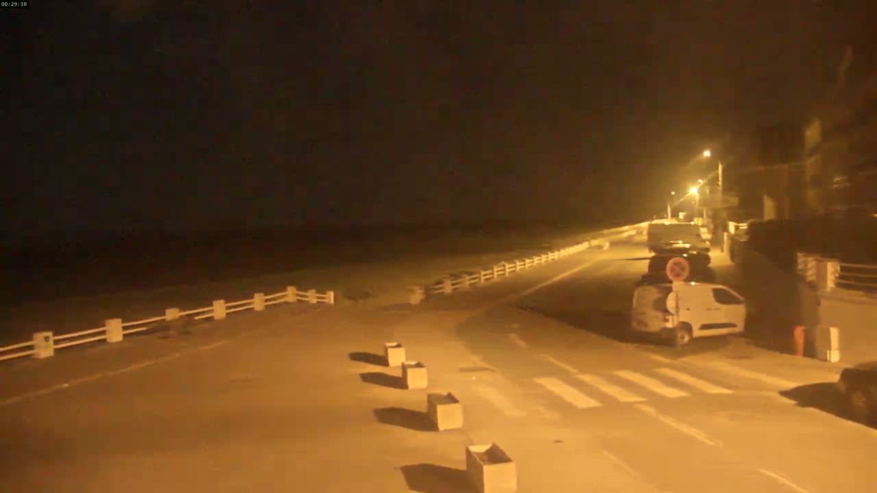 A nighttime street scene shows a road next to a beach, with several vehicles parked along the side, under dimly lit conditions.