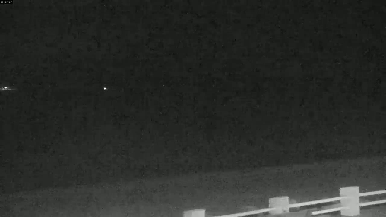 A nighttime view shows two distant lights and a low, partially visible fence in the foreground under a dark, clear sky.