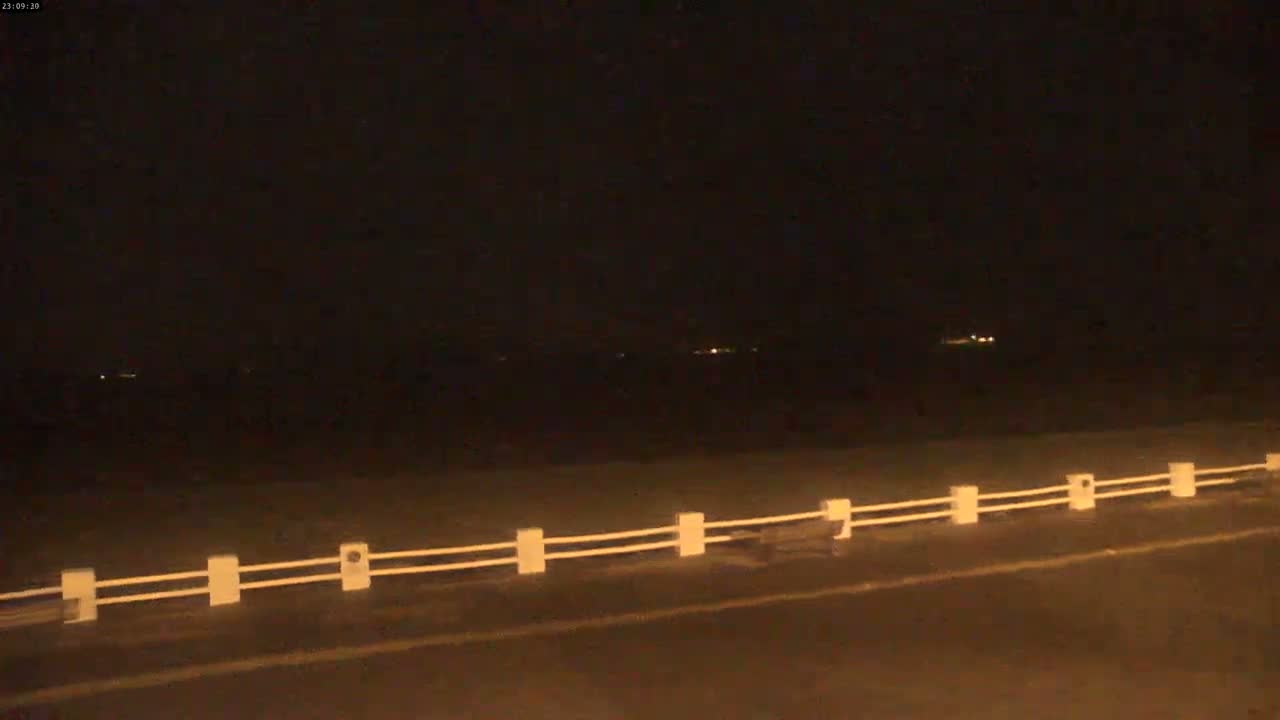 A nighttime view shows a dark body of water with faint lights in the distance, a short section of road with a low white fence running along the edge.