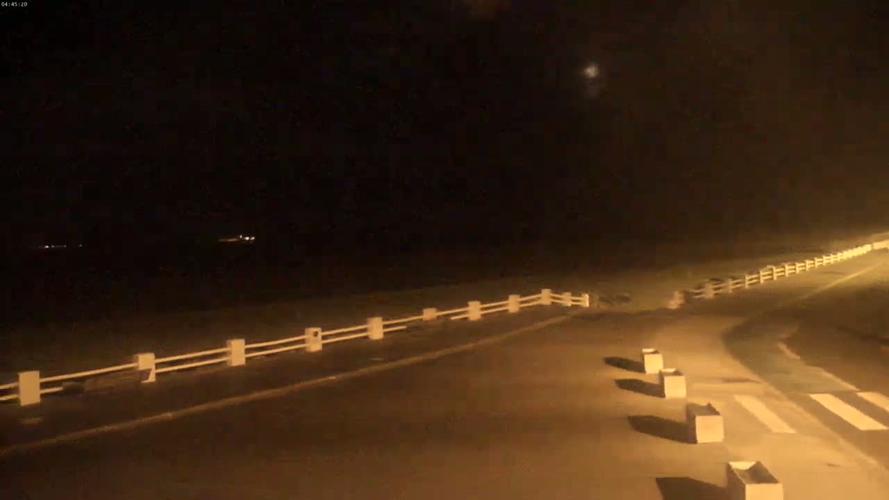 A nighttime view of a paved road with a white fence and several square objects lining the side, under a dark sky.