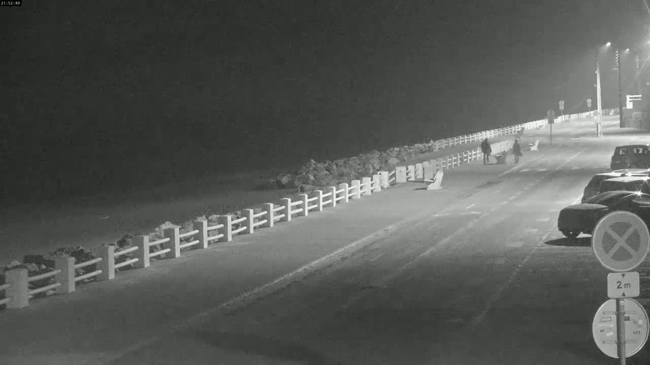 A nighttime scene shows a paved road next to a low white fence bordering a rocky shoreline, with several cars parked along the road and two pedestrians walking.