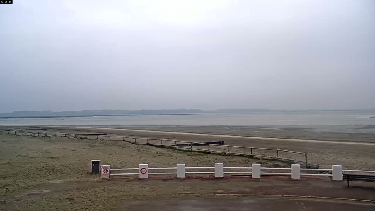 A wide, flat expanse of sand and shallow, calm water stretches to a hazy horizon under an overcast sky.