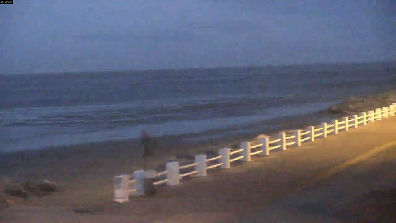 A person walks along a sandy beach at dusk, next to a white fence separating the beach from a paved area, with a dark, somewhat choppy sea in the background.