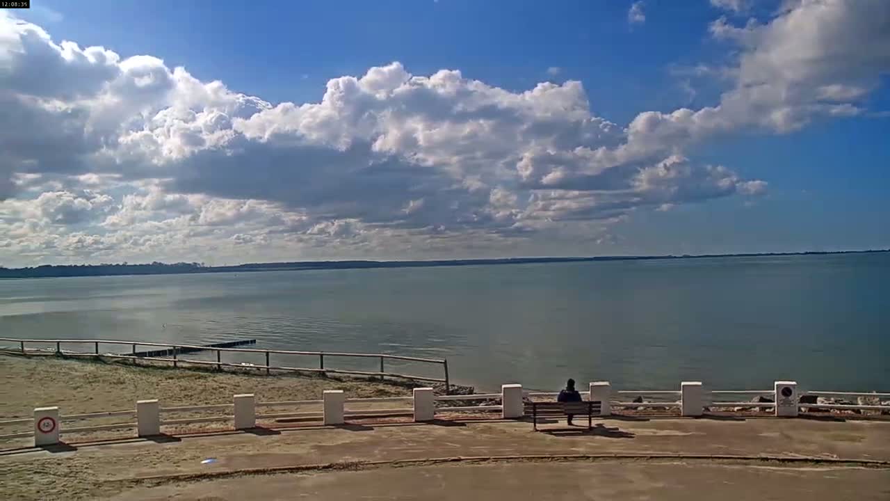 A person sits on a bench overlooking a calm body of water under a partly cloudy sky.