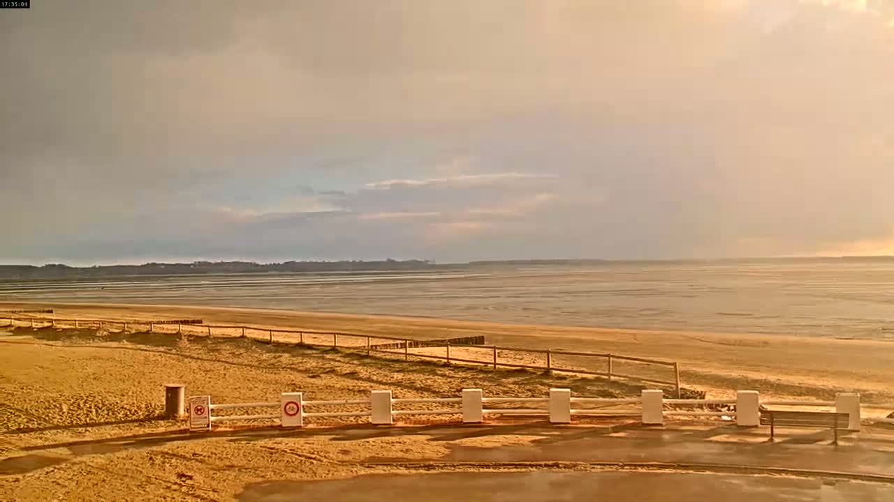 A sandy beach stretches to a calm, wide body of water under a cloudy, pale-gold sky.