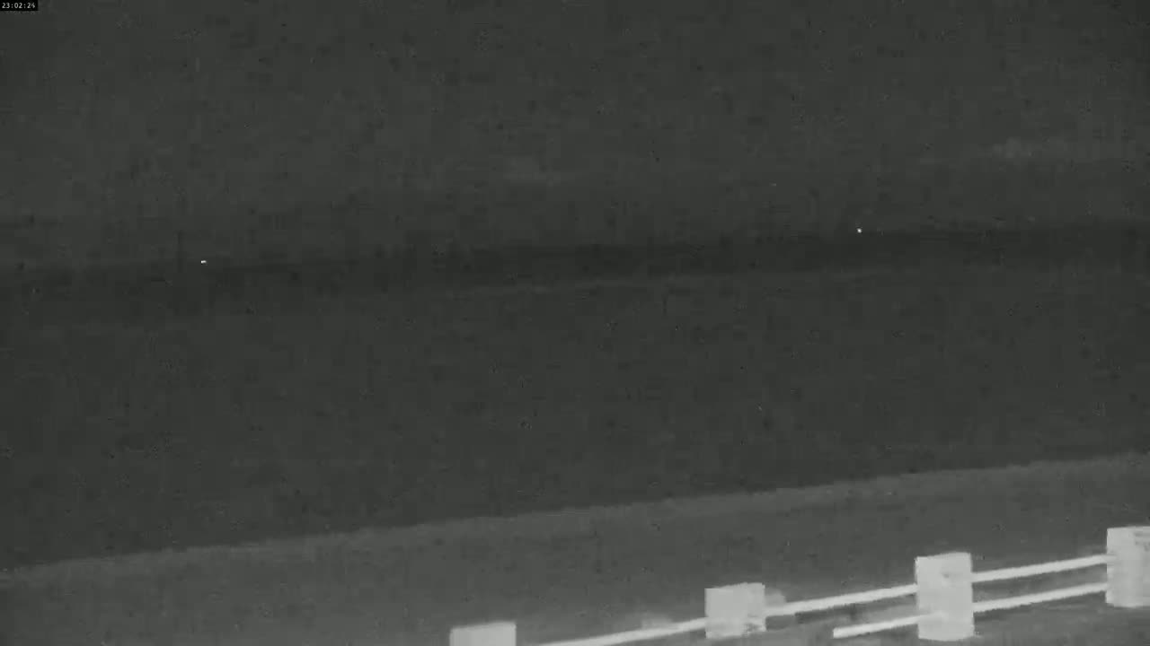 A nighttime, low-light image shows a dark body of water extending to a dark horizon, with a short section of low fence in the foreground.