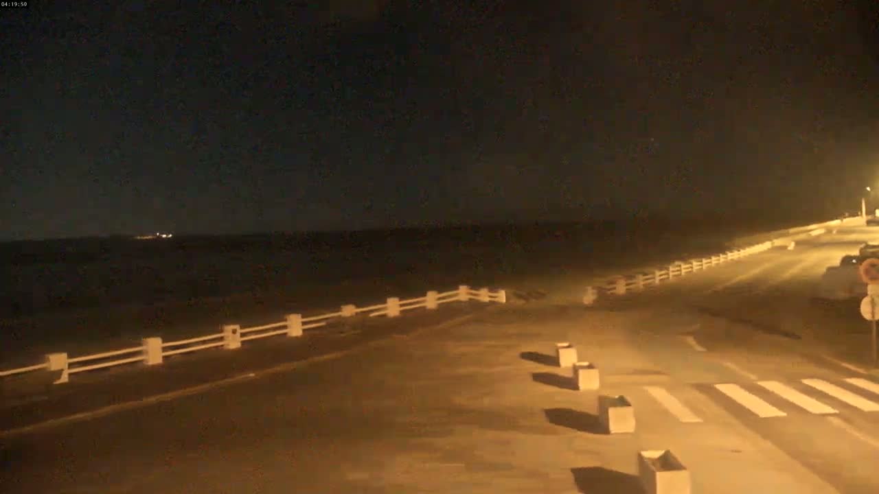 A nighttime view of a paved road next to a body of water, separated by a low white fence, under a dark sky.