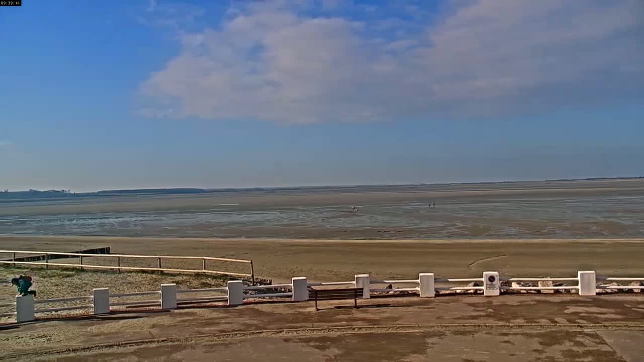 A mostly sunny day shows a wide expanse of flat, muddy beach extending to a calm body of water under a partly cloudy sky.