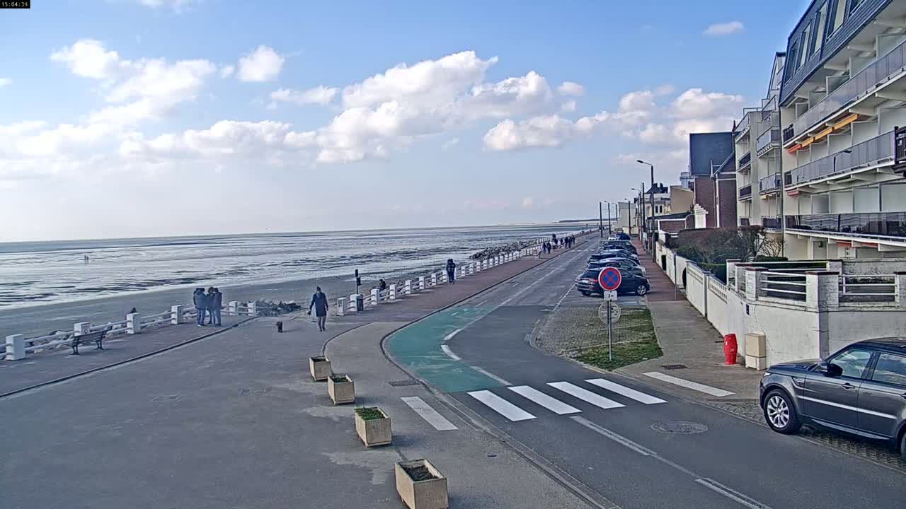 A paved walkway and road next to a beach with exposed mudflats under a partly cloudy sky lead to apartment buildings.