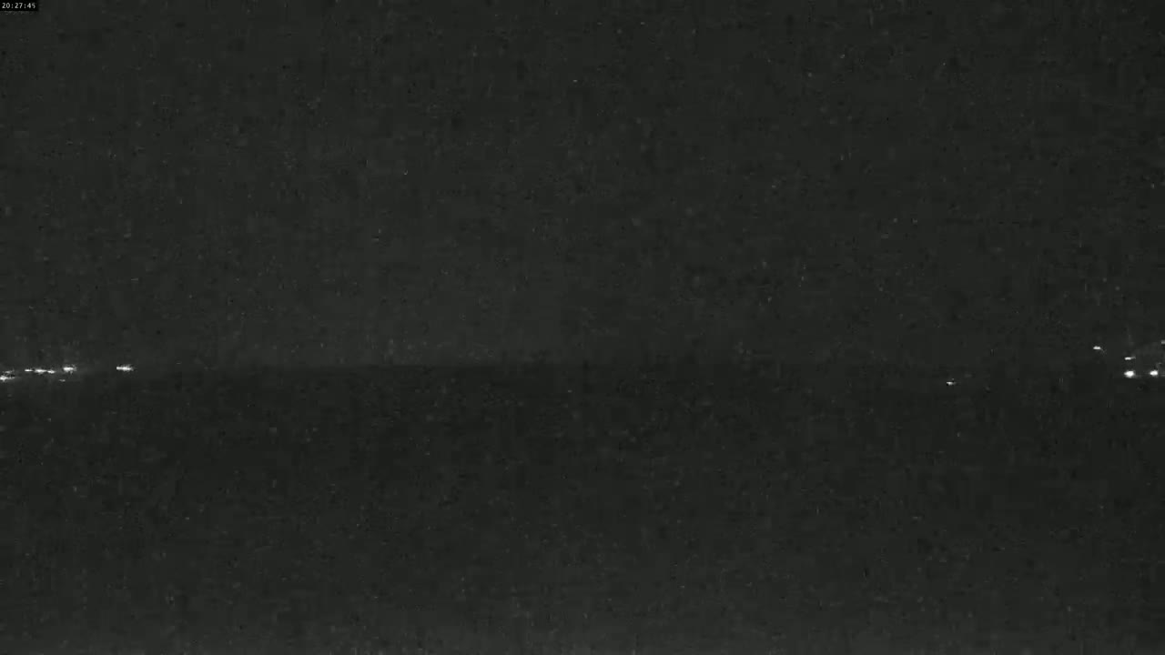 A nighttime, dark view shows a few distant lights on a flat horizon under a mostly clear sky.