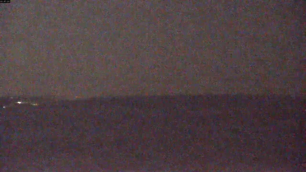 A dark, nighttime scene shows a flat expanse of land or water under a mostly clear, dark sky with a few distant lights visible on the horizon.