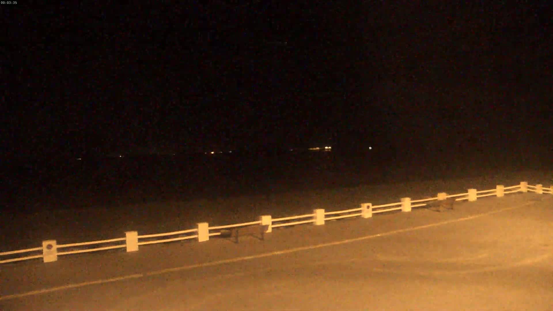 A nighttime view of a road with a white fence, dimly lit by distant lights under a dark sky.