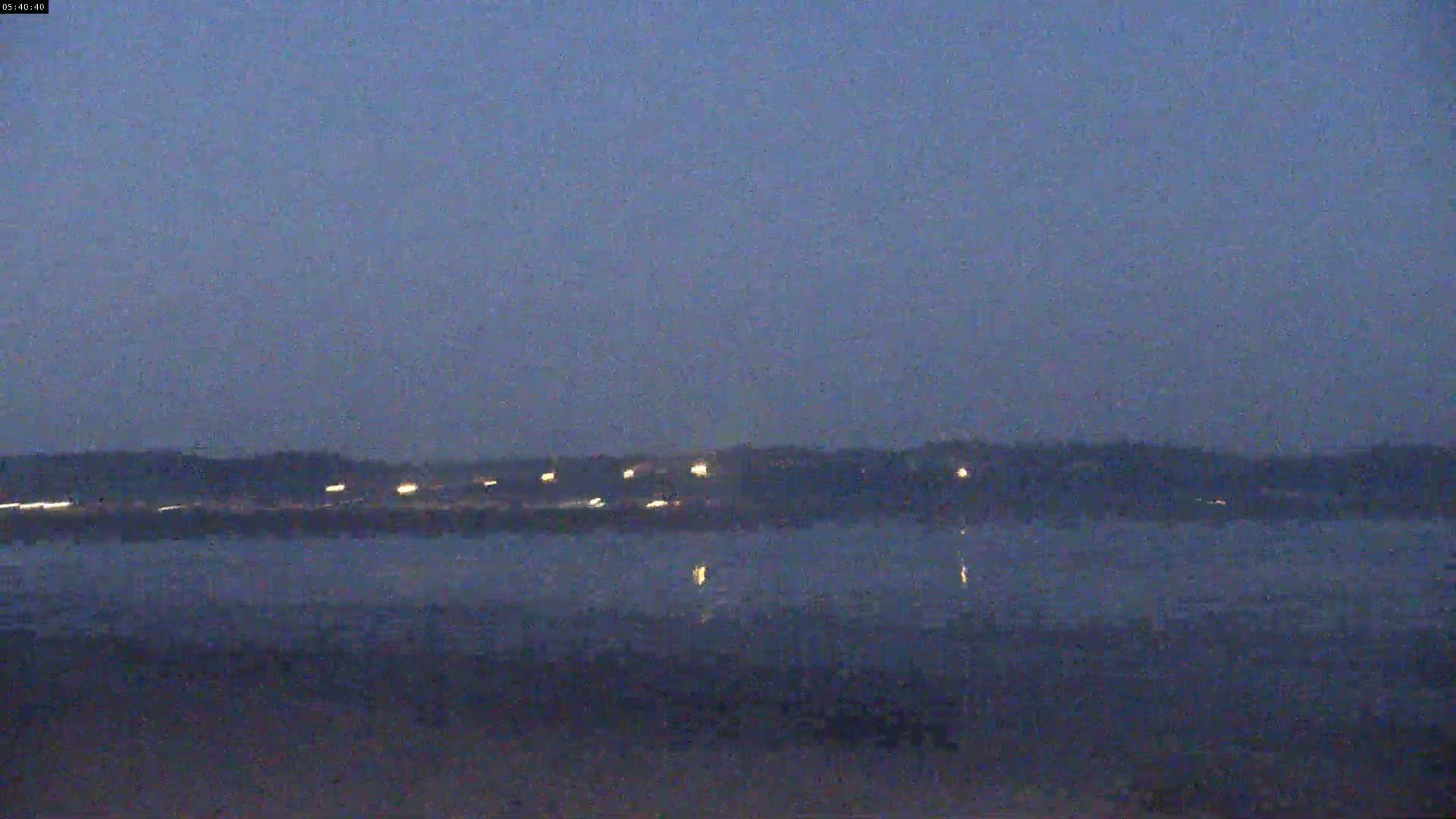 A dark, calm body of water reflects the faint lights of a distant shoreline under a twilight sky.