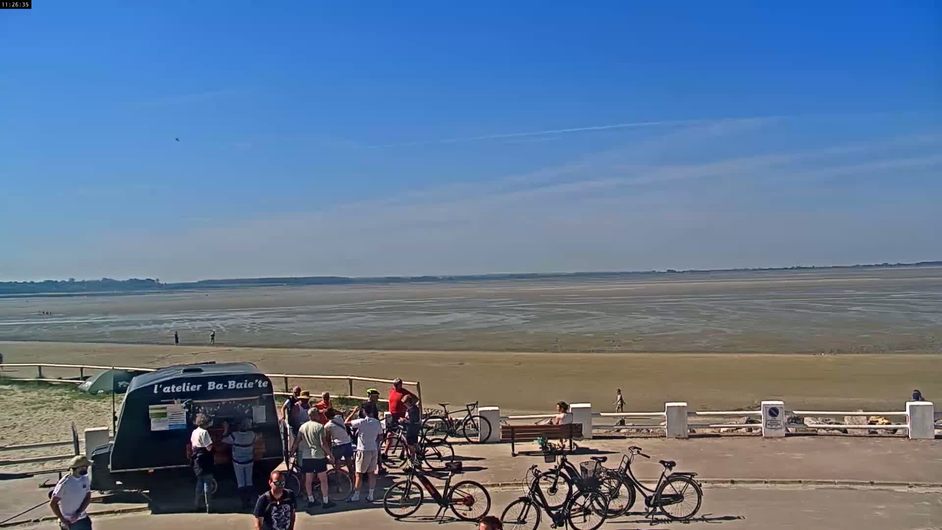 A sunny day at a seaside location shows a group of people gathered near a food trailer and several bicycles, overlooking a wide expanse of exposed sand and mudflats under a clear blue sky.
