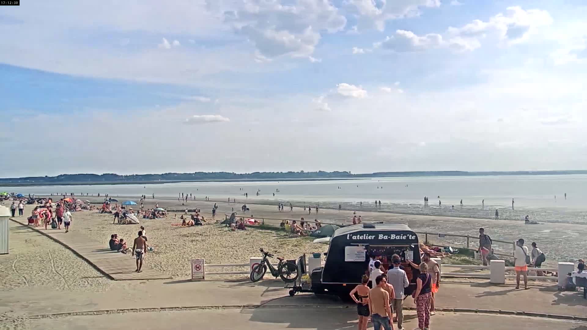 A sandy beach with many people sunbathing and swimming, a food truck, and a calm, partly cloudy sky.