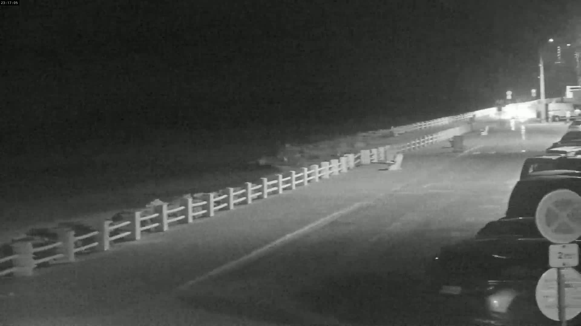 A nighttime, low-light view shows a paved area with several cars parked beside a white fence bordering a dark expanse, possibly a beach or water.