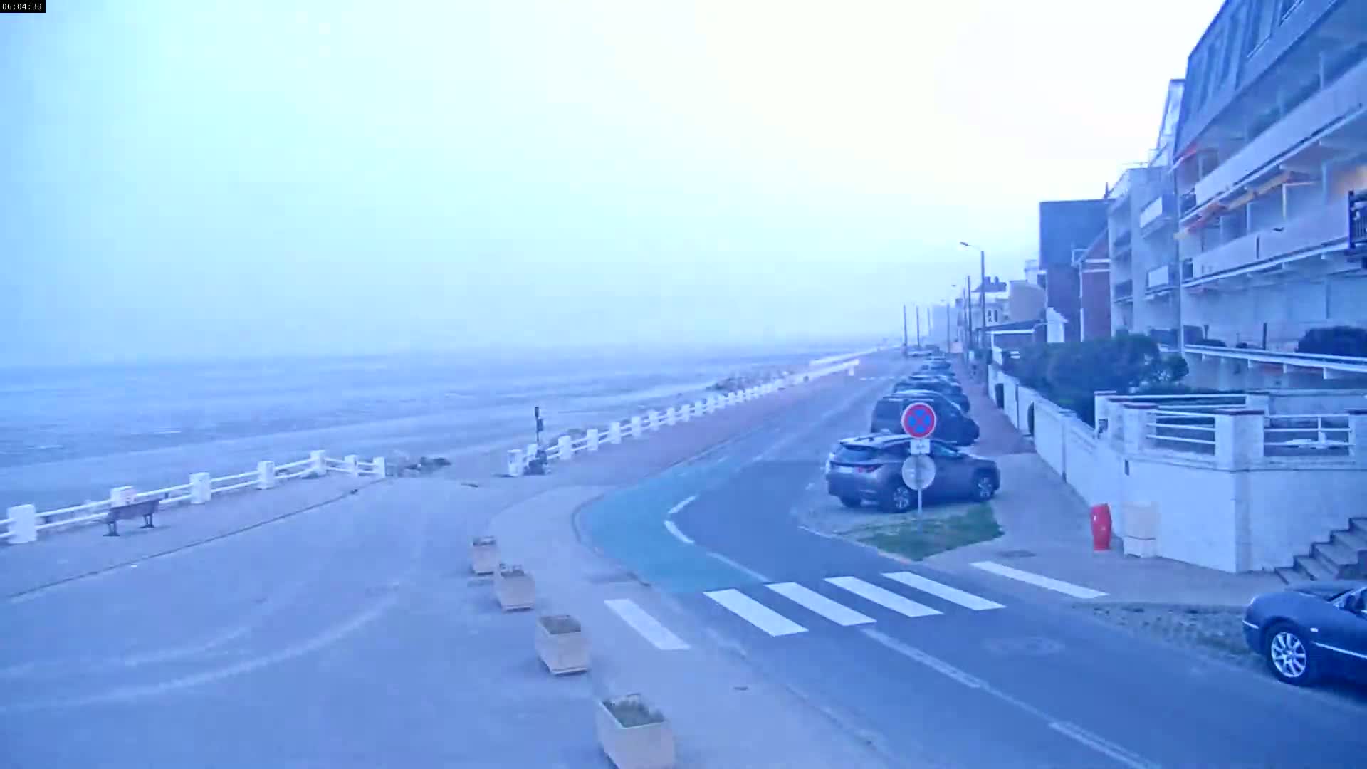 A hazy, overcast day reveals a seaside road with parked cars alongside buildings, leading to a calm, light-grey ocean.