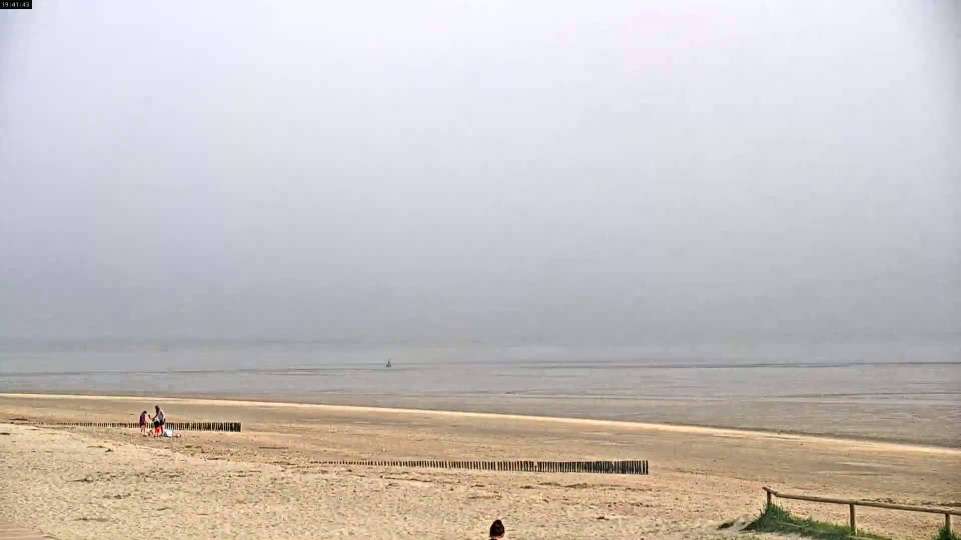A foggy beach scene shows a sandy shore with a few people and a calm, wide body of water extending to the horizon.
