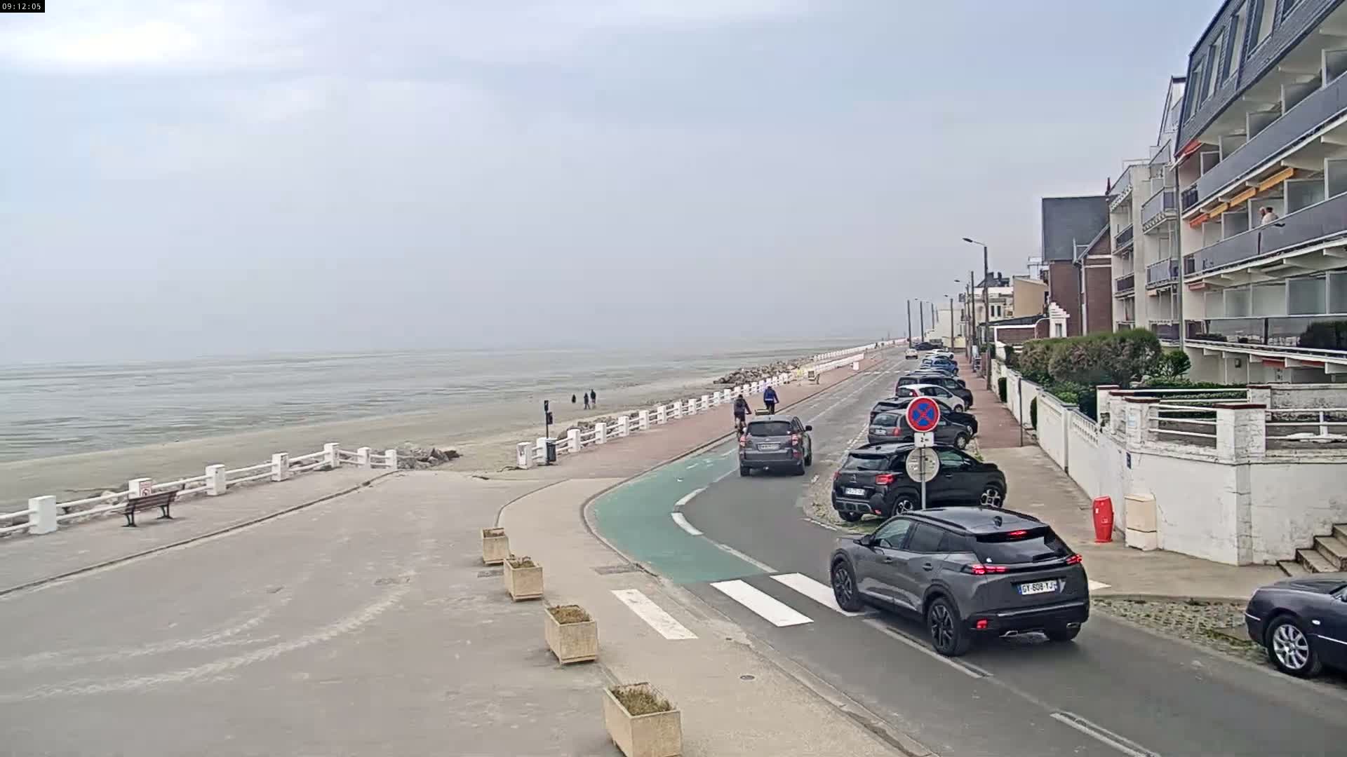 A paved road winds along a beach with parked cars and a few pedestrians and cyclists, under an overcast sky.
