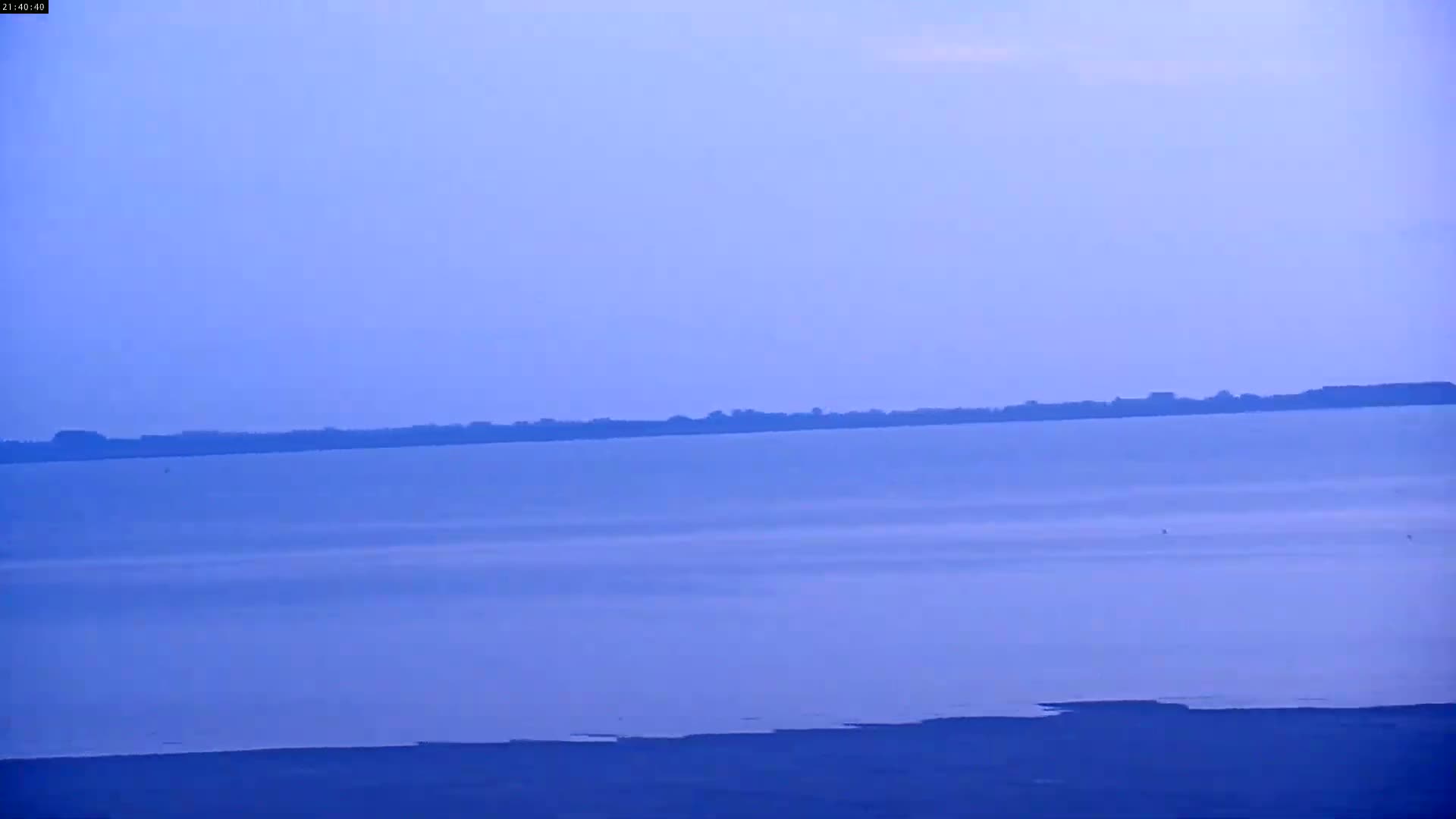 A calm, pale blue body of water stretches to a distant, dark shoreline under a clear, light blue sky.