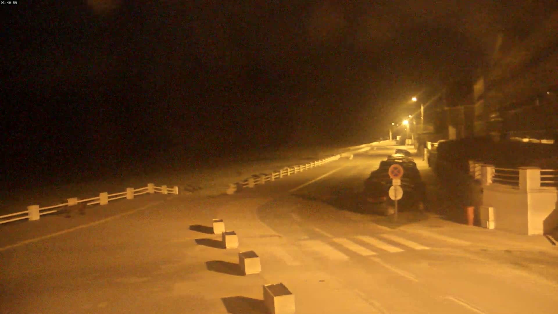 A nighttime view of a road next to a beach, illuminated by streetlights, shows parked cars and a white fence separating the road from the beach.