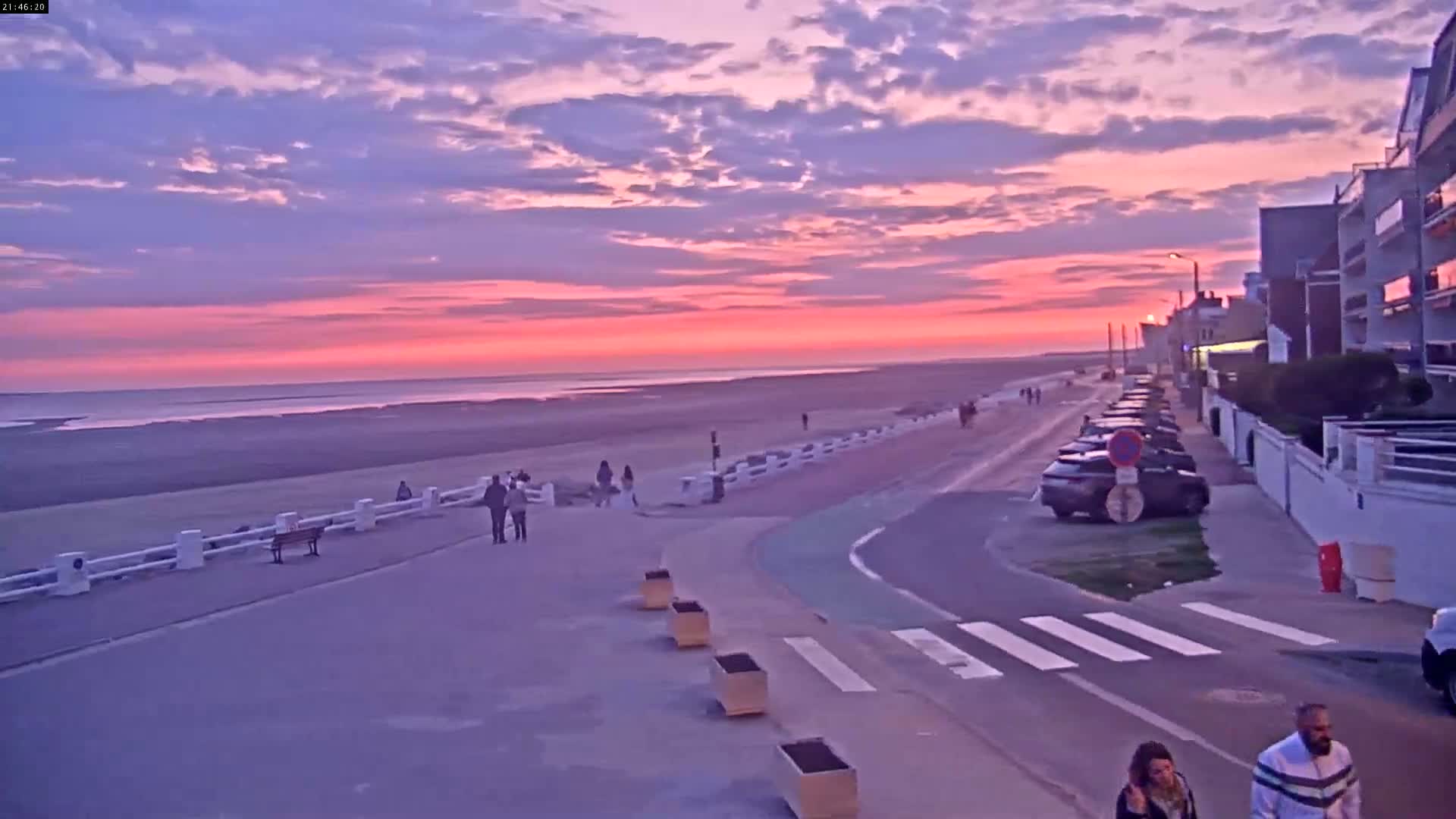 A pink and purple sunset over a beach, with people walking along a paved path next to parked cars and buildings.