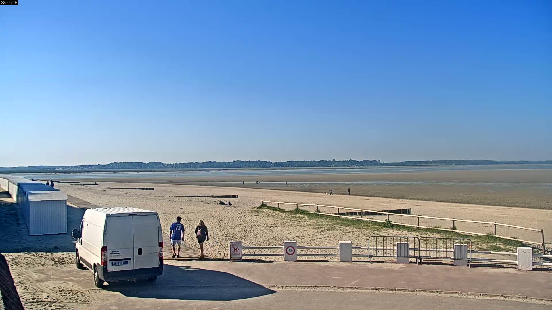 A white van is parked near a sandy beach on a sunny day, with a couple walking toward the water and a line of beach huts visible in the distance.